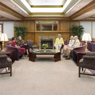 A cozy living room with people sitting and conversing on sofas around a central table, surrounded by warm lighting and plants.