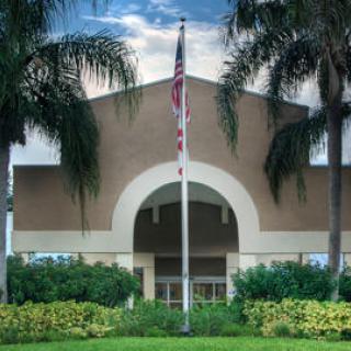 A building with an American flag in front, surrounded by palm trees and lush landscaping.