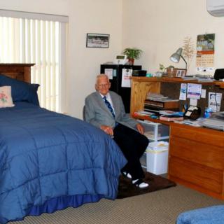 An elderly man sitting at a desk in a bedroom with a blue bedspread.