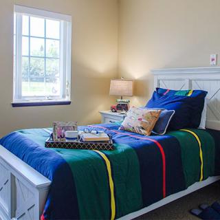 A cozy bedroom with a colorful bedspread, illuminated by a bedside lamp and natural light from the window.