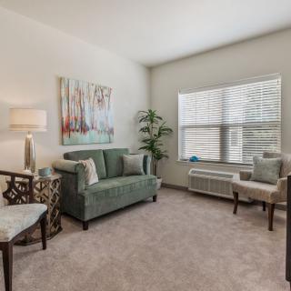 A cozy living room with a sofa, chair, coffee table, and indoor plant, featuring neutral decor and natural light from a large window.