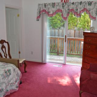 A cozy bedroom with pink carpet and classic furniture, featuring a large window with floral curtains that opens to a view of the garden.