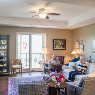 A cozy living room with two people relaxing on sofas, surrounded by home decor and illuminated by natural light from a large window.