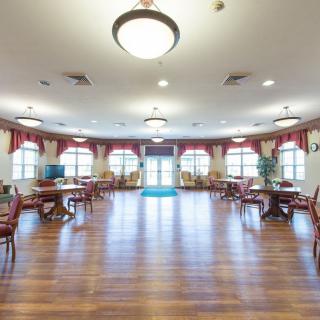 A spacious dining area with wooden flooring, featuring several tables and chairs, well-lit by ceiling lights and natural light from large windows.