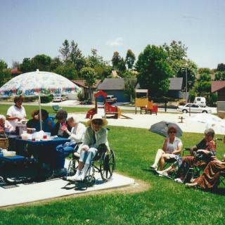 A group of people enjoying a sunny day at a park, sitting together under a large umbrella with some in wheelchairs.