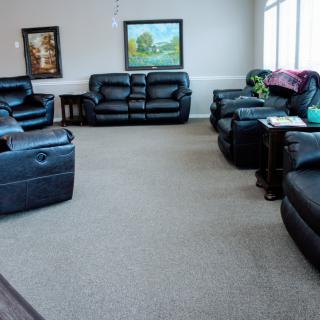 A cozy living room with multiple leather sofas and armchairs arranged around a carpeted floor, complemented by artwork on the walls and large windows for natural light.