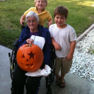 An elderly woman in a wheelchair, holding a carved pumpkin, is accompanied by two smiling children outdoors.