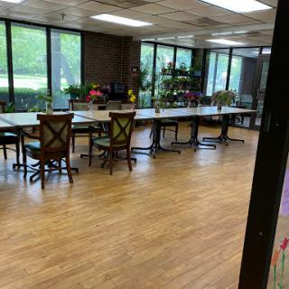 A spacious and well-lit office workspace featuring multiple tables and chairs, surrounded by large windows and greenery.