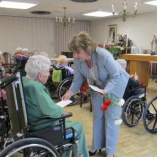 A woman interacts with seniors in wheelchairs at a nursing home, offering them flowers.
