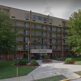 A multi-story residential apartment building with an entrance surrounded by trees and landscaped areas.
