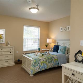 A cozy bedroom featuring a bed with colorful bedding, a dresser, and a lamp by the window.