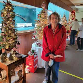 A woman in a red coat stands among festive Christmas decorations and trees indoors.