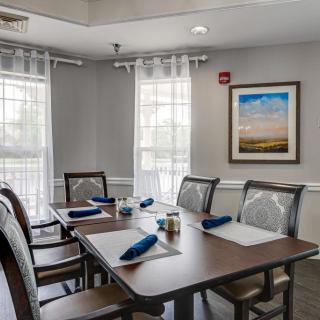 A neatly arranged dining room with a table set for four, featuring blue napkins and placemats, with sunlight streaming through the windows.