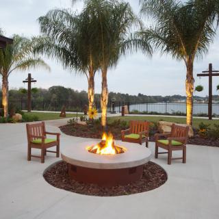 An inviting outdoor area with a fire pit surrounded by chairs, overlooking a scenic lake and framed by palm trees.