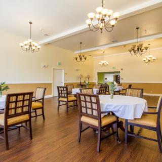 A well-lit dining room with elegantly set tables and chairs, featuring chandeliers and wooden flooring.