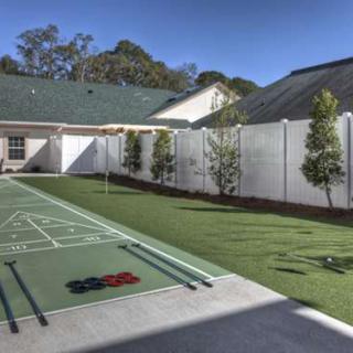A backyard with a shuffleboard court, surrounded by a white fence and trees.