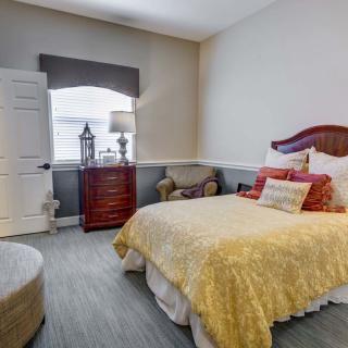A cozy bedroom featuring a bed with a yellow coverlet, a wooden cabinet with a lamp, and an upholstered chair by the window.
