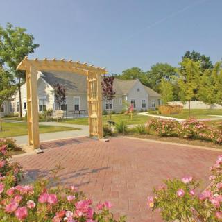 A picturesque garden with a wooden pergola, surrounded by blooming flowers and a paved pathway leading to a charming house.