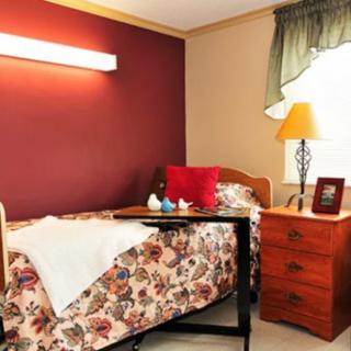 A cozy bedroom with a single bed featuring floral bedding, a nightstand with a lamp, and a warm red accent wall.