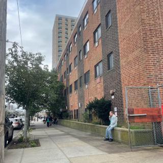 A view of an urban apartment building with a person sitting on the sidewalk, surrounded by greenery and a cloudy sky.