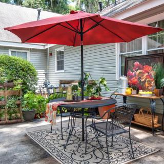 A cozy patio area with a red umbrella, table and chairs, surrounded by lush greenery and potted plants.