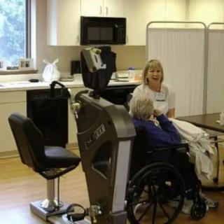 A caregiver assists an elderly person in a wheelchair in a therapy room with medical equipment.