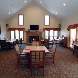A spacious lounge and dining area with a high ceiling, featuring several tables and chairs, large windows, and a central fireplace.