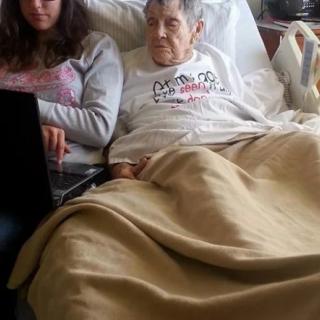 An elderly person resting in bed while a younger person sits nearby using a laptop.