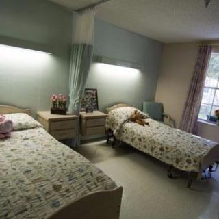 A shared nursing home room with two twin beds, light floral bedding, and a window providing natural light.