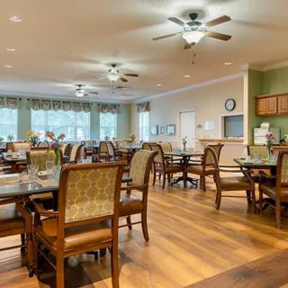 A well-lit dining area with multiple tables and chairs, featuring ceiling fans and large windows.