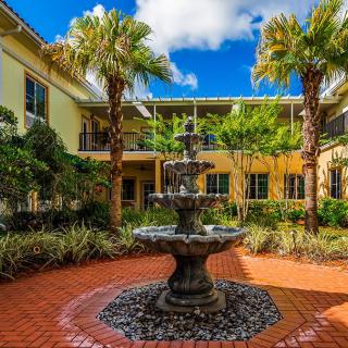 A picturesque courtyard with a central fountain surrounded by palm trees and a charming building under a clear blue sky.