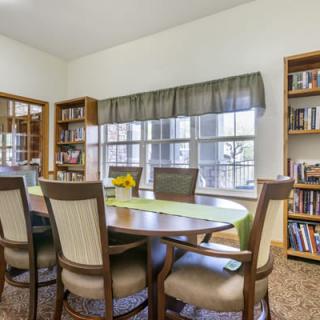 A cozy library or meeting room with a wooden table, surrounded by chairs and bookshelves, lit by natural light from a large window.