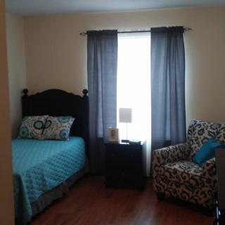 A cozy bedroom featuring a single bed, an armchair, and a window with blue curtains.