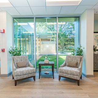 A cozy waiting area featuring two stylish chairs, a small table, and plenty of natural light from large windows.