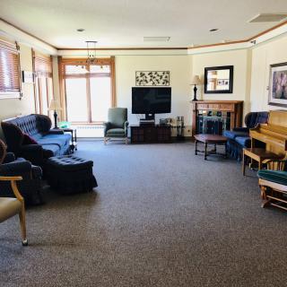 A spacious living room with a television, sofa, piano, and chairs arranged around a central carpeted area.