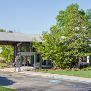 A modern building entrance surrounded by trees and a parking lot during the day.