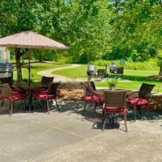 A peaceful outdoor patio with tables, chairs, and an umbrella, surrounded by lush greenery.