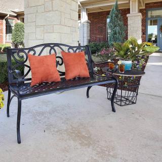 A wrought iron bench with orange pillows on a patio surrounded by garden plants.