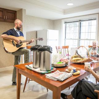 A casual gathering in a kitchen setting, with one person playing the guitar while another enjoys a meal.