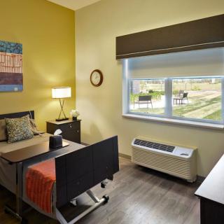 A well-lit hospital room with a single bed, side table, and a large window overlooking green fields.