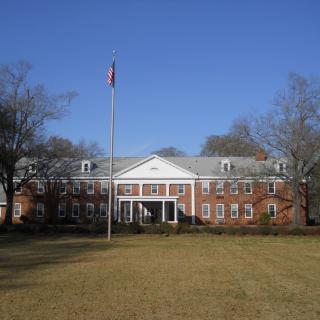 A red brick building with a flagpole in front, surrounded by trees and grass on a sunny day.