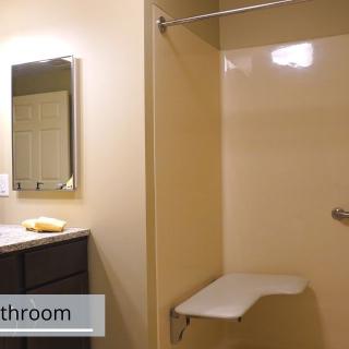 An accessible bathroom with a sink, mirror, and shower area featuring a folding seat.