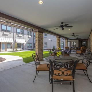 A covered outdoor patio area with seating, overlooking a landscaped garden and building exterior.