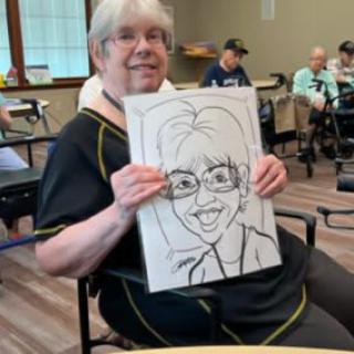 An elderly woman proudly holds up a caricature drawing of herself in a community setting.