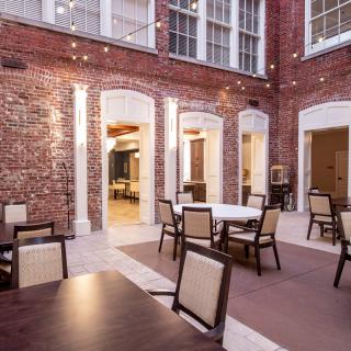 A cozy courtyard with tables and chairs, surrounded by brick walls and illuminated by string lights.