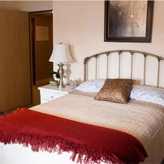 A cozy bedroom with a neatly made bed featuring pillows and a red blanket, a nightstand with a lamp, and artwork on the wall.