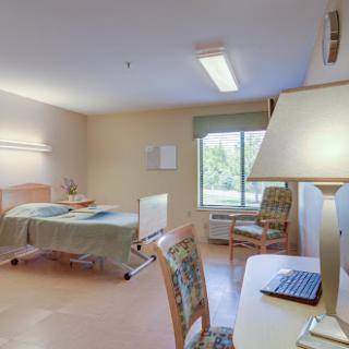 A well-lit hospital patient room featuring a bed, chairs, and a desk with a computer.