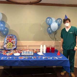 A festive birthday setup featuring blue decorations, a cake, and a person in medical scrubs celebrating.