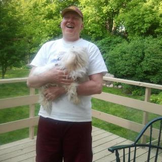 A person smiling and holding a dog on a wooden deck surrounded by greenery.