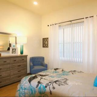 A cozy bedroom with a bed covered in a patterned comforter, a dresser with a lamp and mirror, and a blue armchair by the window.